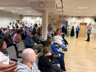 Duża grupa ludzi siedzi i stoi w sali z jasną, beżowo-zieloną, wzorzystą ścianą. Mężczyzna po prawej stoi z mikrofonem i przemawia. Publiczność siedzi w rzędach i przy ścianie, część osób stoi z tyłu. W pomieszczeniu jest jasna drewniana podłoga i sufit z lampami, na środku sufitu wisi zegar.