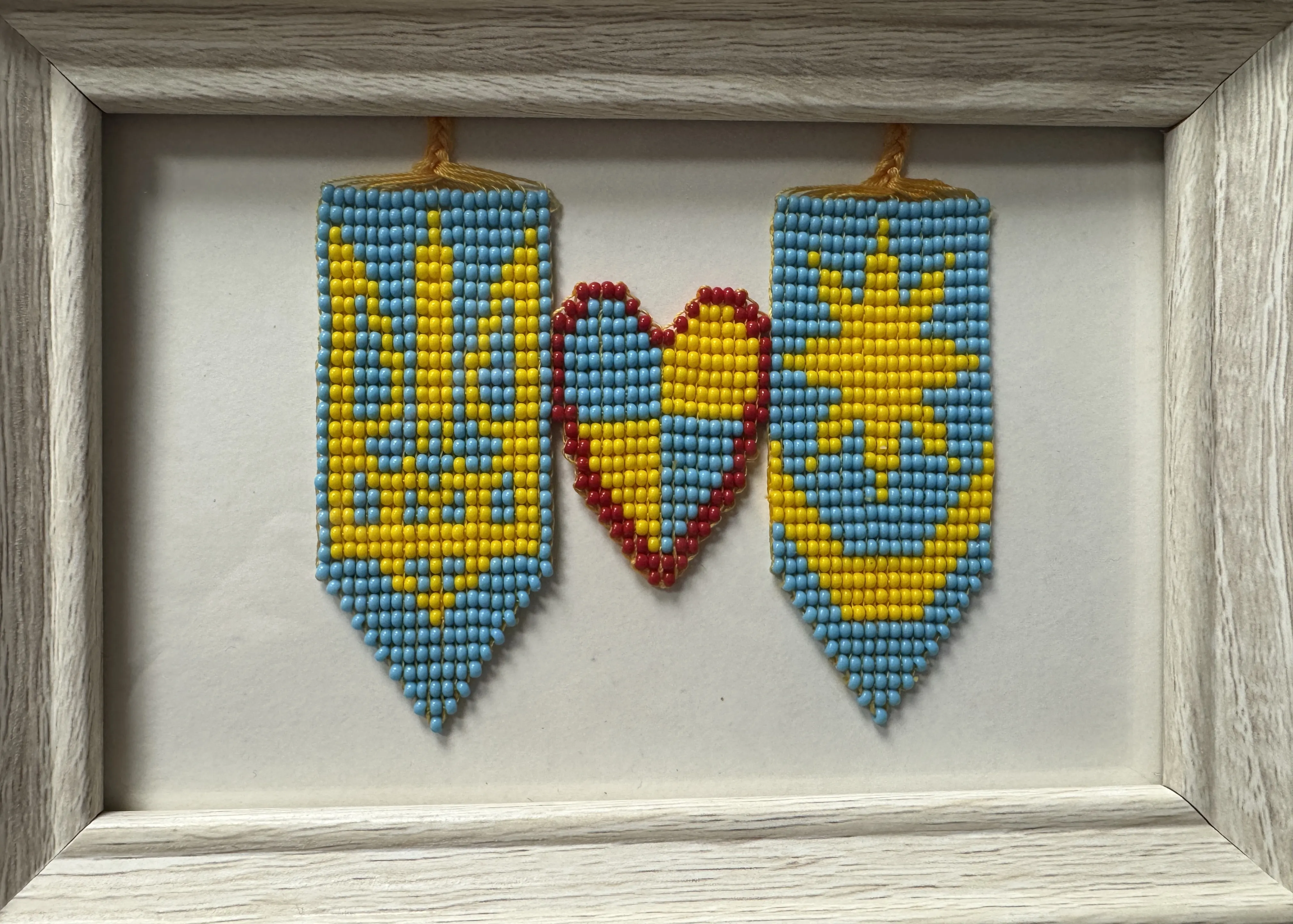 Trzy zawieszone ozdoby z małych koralików na białym tle w jasnej drewnianej ramie: po bokach dwie niebiesko‑żółte chorągiewki z zakończeniem w szpic, pośrodku serce wypełnione na przemian niebieskimi i żółtymi koralikami obramowane czerwonymi.