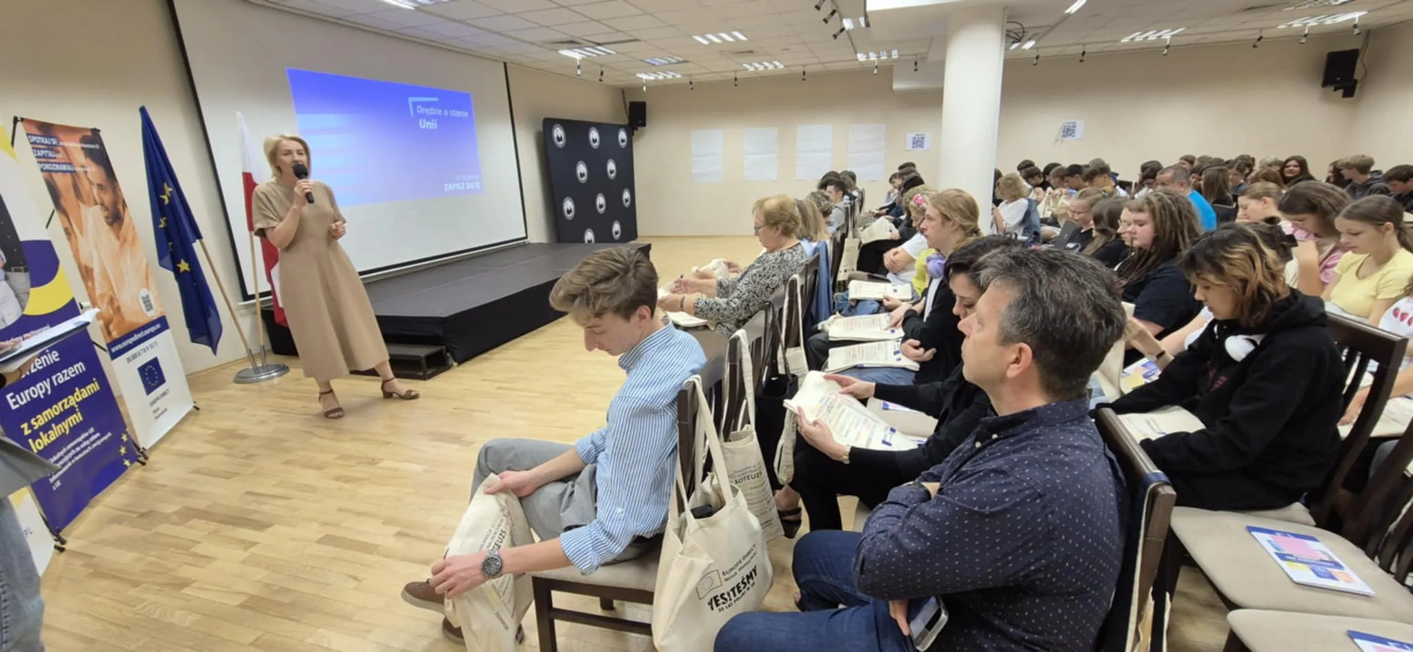 Kobieta stoi i przemawia do około czterdziestu osób siedzących na krzesłach w sali konferencyjnej z jasnymi ścianami i drewnianą podłogą. Po lewej stronie widać flagi Polski i Unii Europejskiej oraz plansze z napisami. Uczestnicy trzymają materiały edukacyjne i bawełniane torby z napisem.