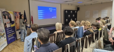 Sala konferencyjna z kilkudziesięcioma osobami siedzącymi w rzędach na drewnianych krzesłach. Po lewej stronie stoi kobieta mówiąca do mikrofonu, obok niej trzy stojące banery i flagi. Na środku ściany znajduje się duży ekran z niebieskim slajdem prezentacji. Pomieszczenie jest jasne, oświetlone sztucznym światłem.