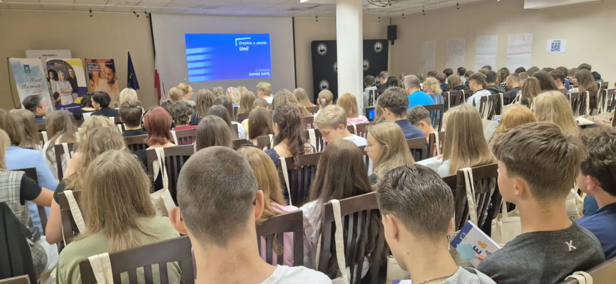 Wielu młodych ludzi siedzi na krzesłach w sali konferencyjnej skierowanych w stronę ekranu z projekcją. W tle wiszą flagi i stoją plansze informacyjne. Sala jest jasna i pełna uczestników.