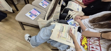 Kilka osób siedzi na krzesłach w sali, trzymając na kolanach bawełniane torby z kolorowym nadrukiem i książeczki. Podłoga drewniana, wnętrze jasne.