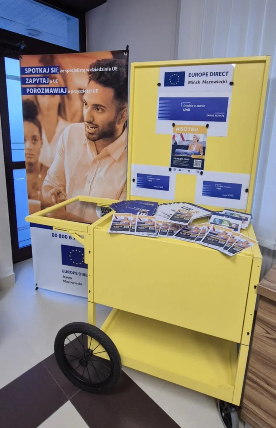 Stoisko informacyjne Europe Direct Mińsk Mazowiecki z żółtym wózkiem, na którym leżą ulotki i broszury. Za nim stoi plakat z wizerunkiem mężczyzny oraz tekstem zachęcającym do spotkań i rozmów o Unii Europejskiej. W tle widać częściowo okno i fragment jasnej ściany.