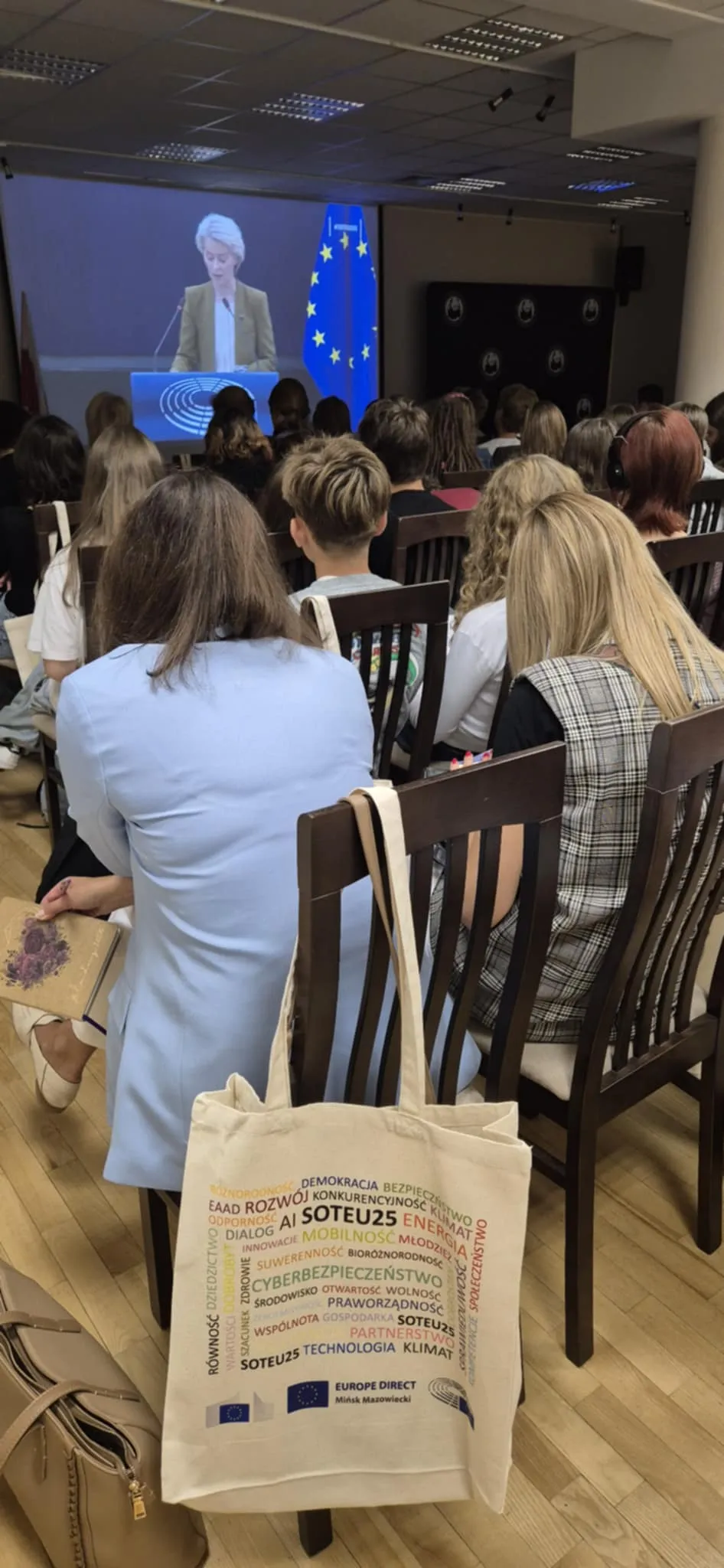Grupa osób siedzi na krzesłach w sali konferencyjnej, oglądając prezentację wyświetlaną na dużym ekranie. Na ekranie kobieta przemawia przy mównicy z symbolem Parlamentu Europejskiego i obok znajduje się flaga Unii Europejskiej. Na pierwszym planie na oparciu krzesła wisi materiałowa torba z kolorowymi napisami dotyczącymi tematyki europejskiej.