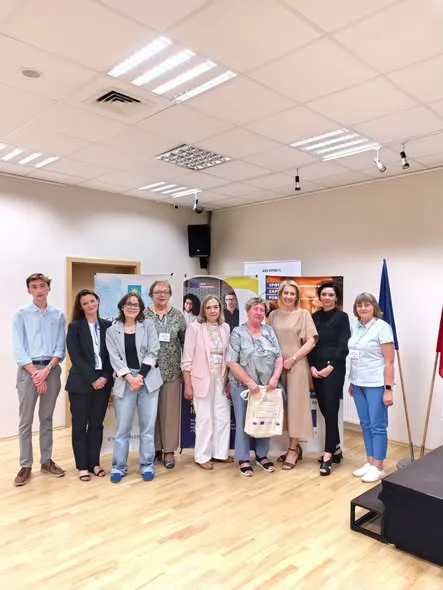 Dziesięć osób stoi w rzędzie w jasnym pomieszczeniu z drewnianą podłogą i sufitowymi panelami świetlnymi. W tle widoczne są dwa stojaki z banerami oraz fragment flagi. Po prawej stronie znajduje się podest.