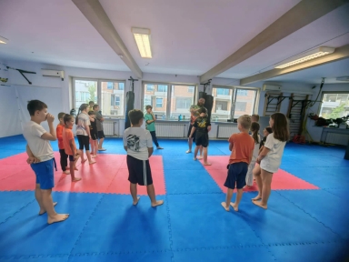 Grupa dzieci w wieku szkolnym stoi na matach do sportów walki podczas zajęć prowadzonych przez instruktora w sali treningowej.