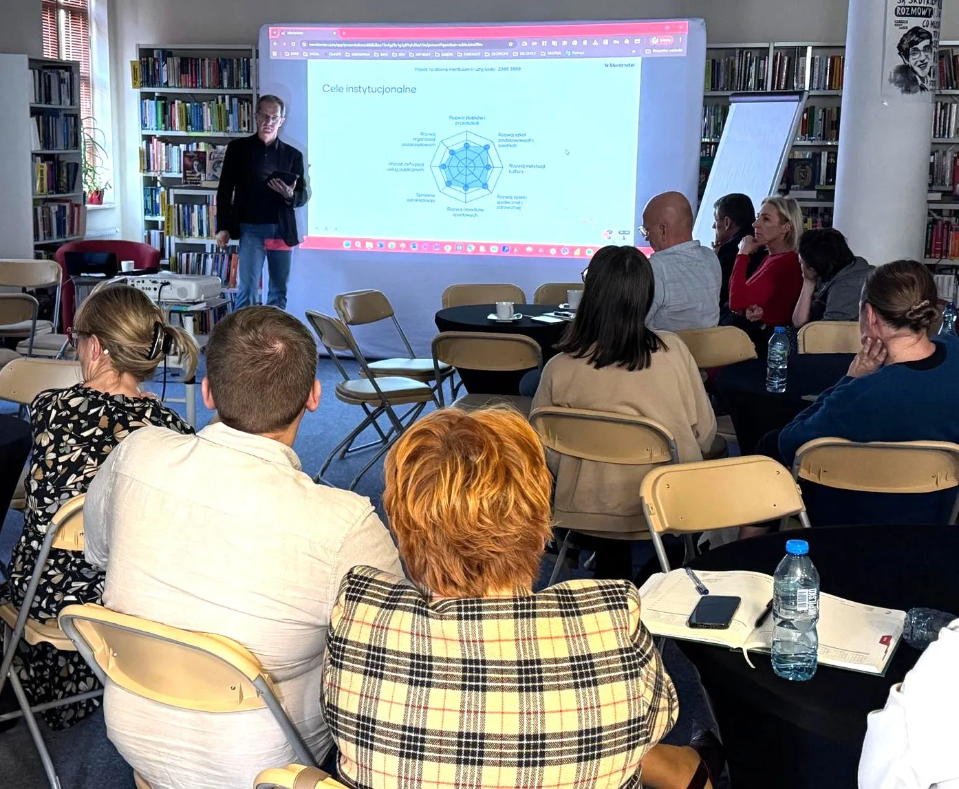 Grupa osób siedzi na składanych krzesłach w sali z regałami z książkami. Mężczyzna stoi przy ekranie z wyświetlaną prezentacją zatytułowaną "Cele instytucjonalne". Na stoliku w pierwszym planie leżą notesy, telefon i butelka wody.