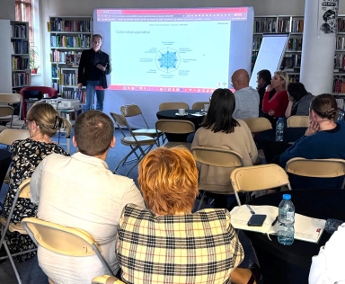 Grupa osób siedzi na składanych krzesłach w sali z regałami z książkami. Mężczyzna stoi przy ekranie z wyświetlaną prezentacją zatytułowaną "Cele instytucjonalne". Na stoliku w pierwszym planie leżą notesy, telefon i butelka wody.