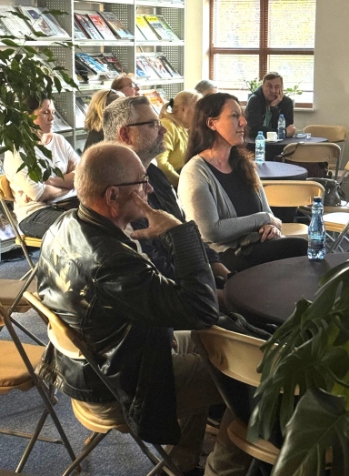 Grupa ośmiu dorosłych osób siedzi na krzesłach i rozmawia w jasnym, dobrze oświetlonym pomieszczeniu z dużym oknem i regałem na czasopisma.