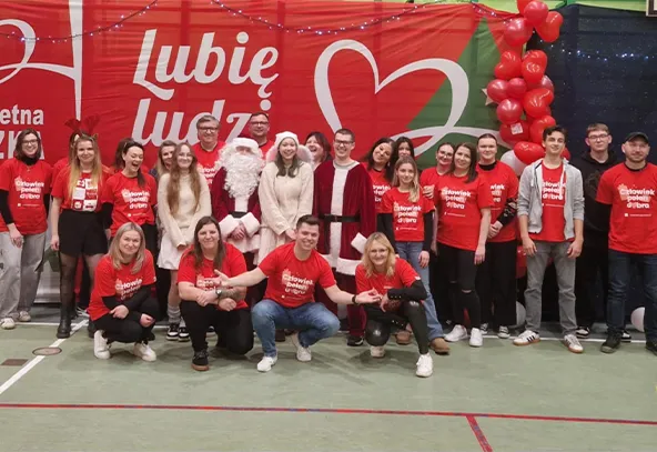 Grupa około dwudziestu osób pozujących na hali sportowej na tle czerwono-zielonego banera z napisem „Lubię ludzi” i sercem. Większość osób nosi czerwone koszulki z białym napisem. W środku stoi osoba przebrana za Świętego Mikołaja oraz kilka osób w białych ubraniach. Po prawej stronie balony czerwone ułożone w kolumnę.