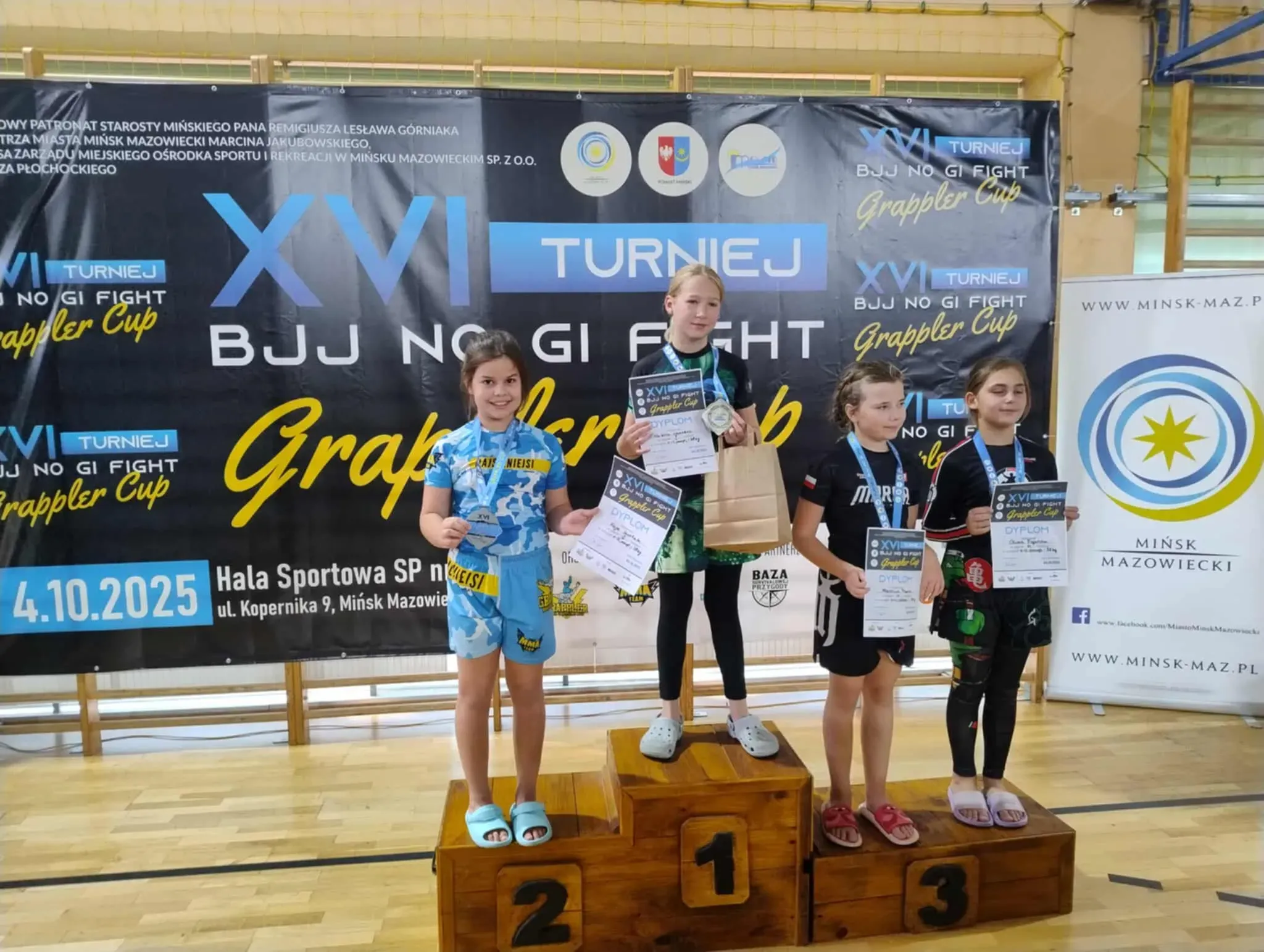 Cztery dziewczynki stoją na drewnianym podium na hali sportowej. Trzy z nich mają medale na szyi i trzymają dyplomy. W tle widoczny jest czarny baner z napisem „XVI TURNIEJ BJJ NO GI FIGHT Grappler Cup”. Po prawej stronie stoi biały roll-up z logo i napisem „Mińsk Mazowiecki”.