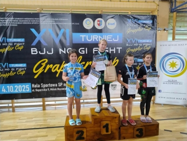 Cztery dziewczynki stoją na drewnianym podium na hali sportowej. Trzy z nich mają medale na szyi i trzymają dyplomy. W tle widoczny jest czarny baner z napisem „XVI TURNIEJ BJJ NO GI FIGHT Grappler Cup”. Po prawej stronie stoi biały roll-up z logo i napisem „Mińsk Mazowiecki”.