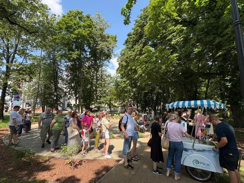 Na zewnątrz. Ludzie stojący w kolejce do saturatora. Wokół drzewa.