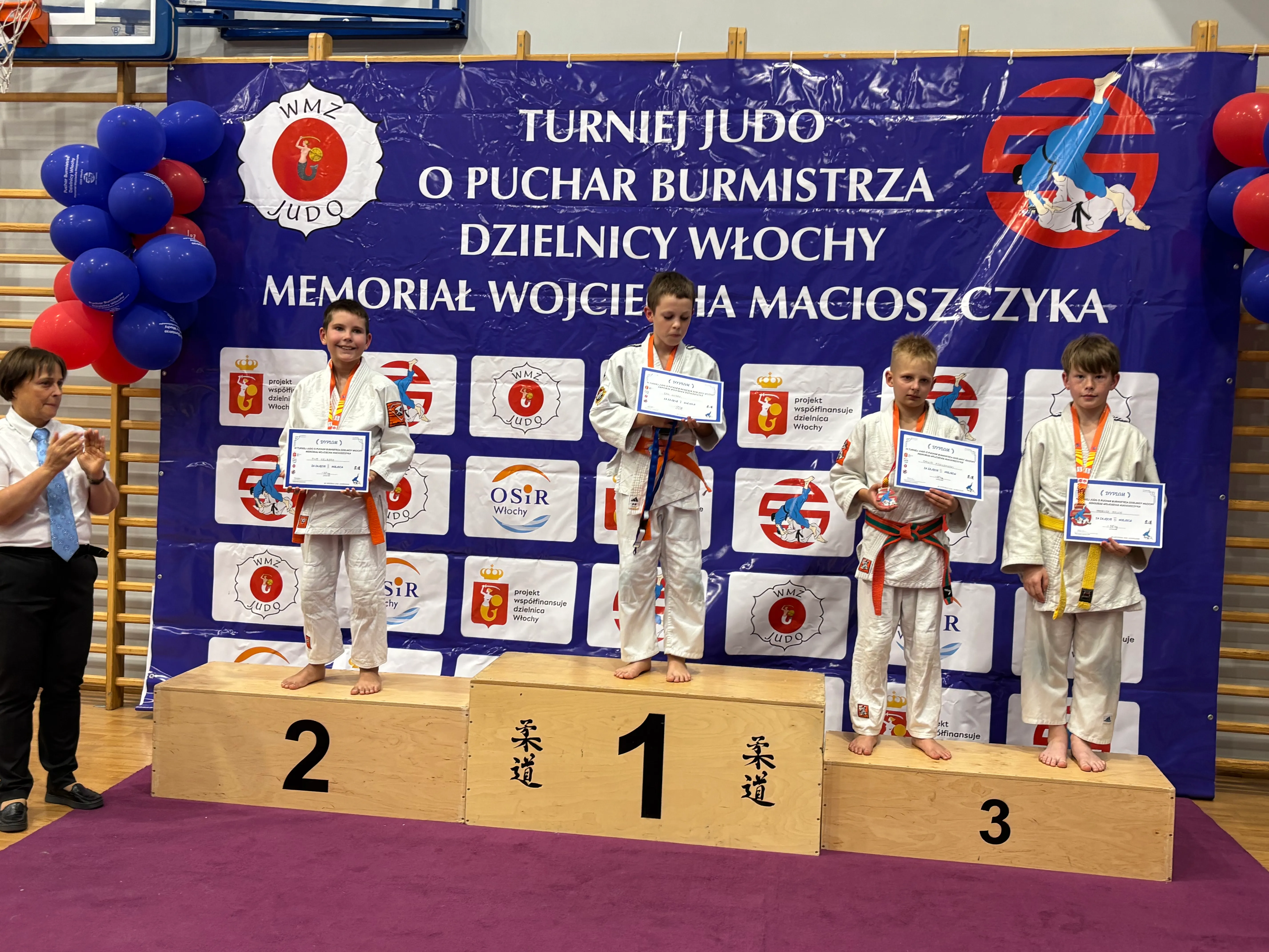 Czterech chłopców w judogach stoi na podium z dyplomami i medalami w hali sportowej podczas turnieju judo.