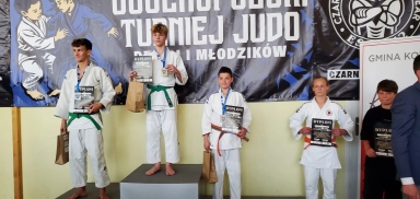 Pięciu zawodników na podium podczas turnieju judo, każdy trzyma dyplom i torbę, w tle baner z napisem o turnieju.
