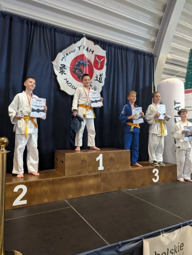 Pięcioro dzieci stoi na podium zawodów judo w hali sportowej. Trzy dzieci na podium mają białe kimona, jedno niebieskie. Wszystkie trzymają dyplomy i uśmiechają się. Za nimi czarna kurtyna i duże godło klubu judo.