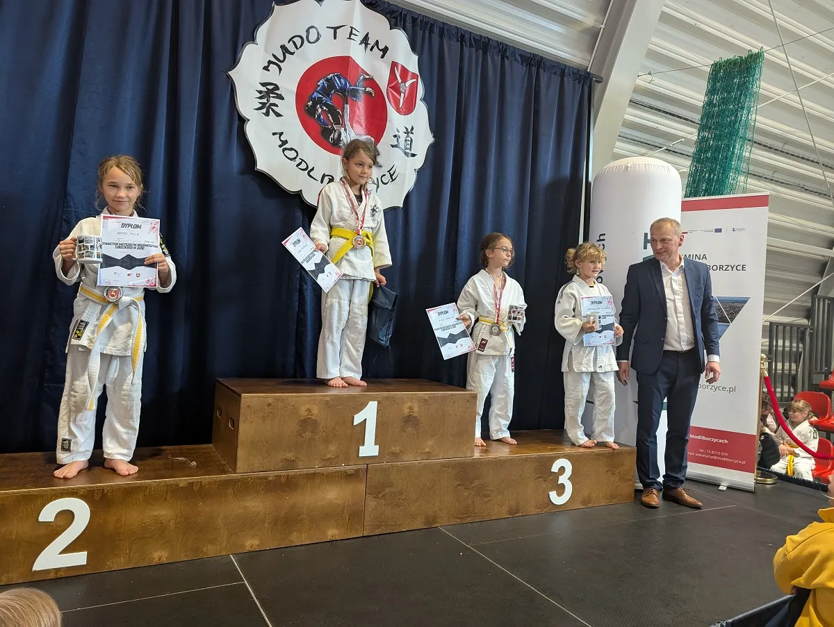 Cztery dziewczynki w białych judogach stoją na podium z trzema stopniami, trzymając dyplomy i medale. Po prawej stoi mężczyzna w garniturze. W tle duży baner z napisem MUDO TEAM MODLICE.