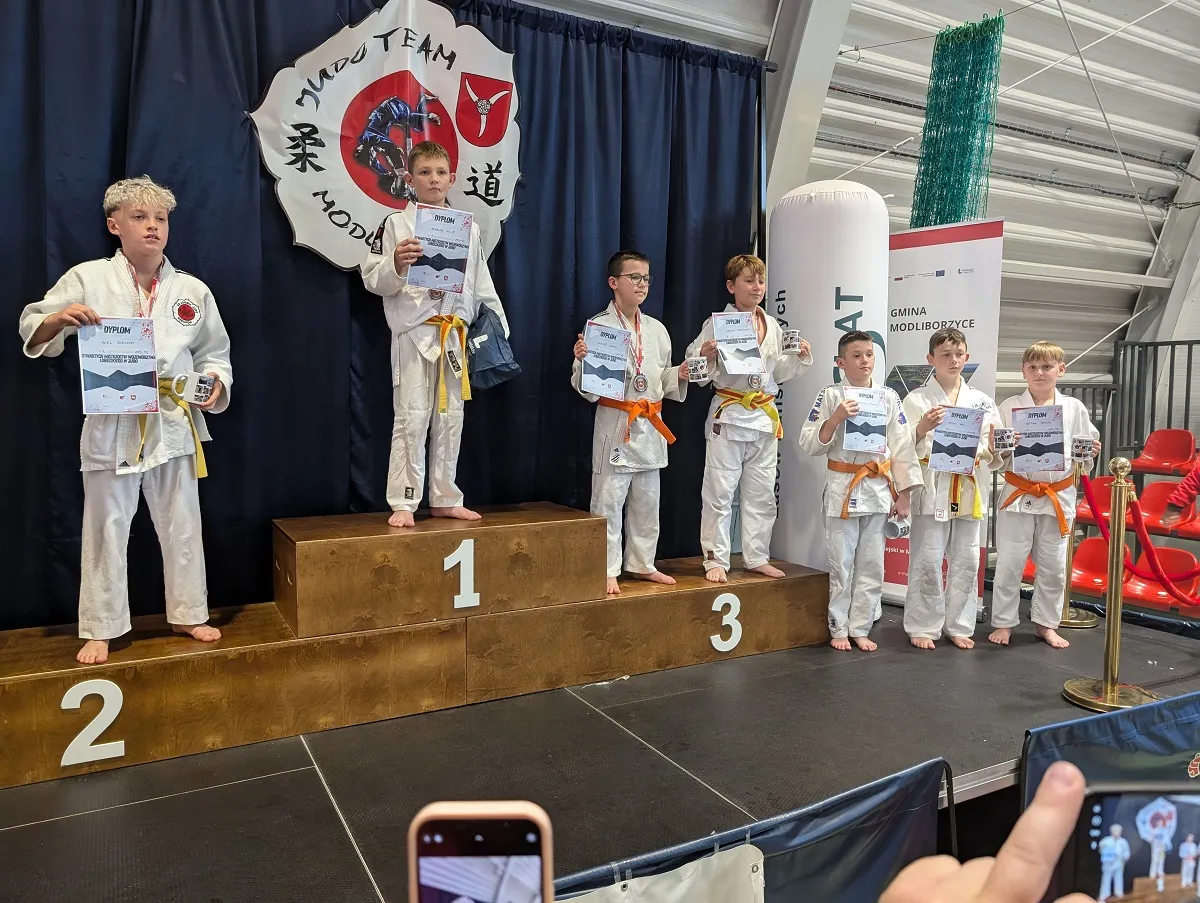 Siedmiu chłopców w białych judogach stoi na podium i obok niego na scenie w hali sportowej. Troje chłopców stoi na podium na miejscach 1, 2 i 3, trzymając dyplomy i kubki. Czterech chłopców stoi obok podium także z dyplomami i kubkami. W tle czarna kurtyna z logo Mudo Team Modliborzyce. Po prawej białe i czerwone roll-upy oraz czerwone krzesła. Przed sceną widać ujęcie dłoni trzymającej telefon.