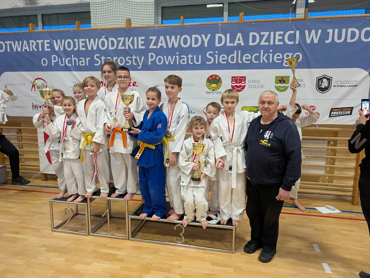 Grupa dzieci w strojach do judo stoi na podium w hali sportowej, trzyma puchary i medale, obok nich stoi mężczyzna w ciemnej bluzie, w tle duży niebieski baner z tekstem o zawodach judo dla dzieci.