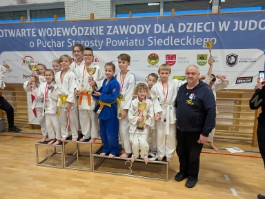Grupa dzieci w strojach do judo stoi na podium w hali sportowej, trzyma puchary i medale, obok nich stoi mężczyzna w ciemnej bluzie, w tle duży niebieski baner z tekstem o zawodach judo dla dzieci.