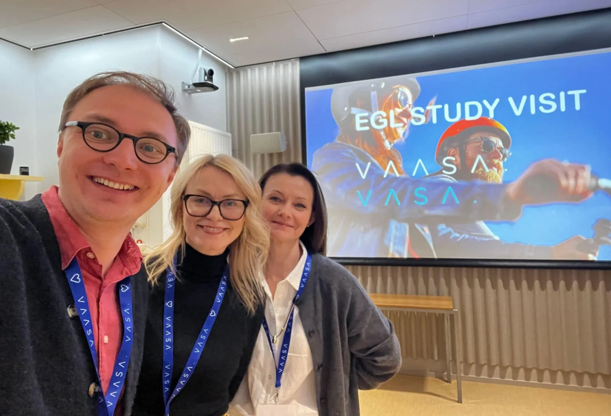 Trzy osoby stoją blisko aparatu i uśmiechają się. Mężczyzna z okularami stoi po lewej. Dwie kobiety stoją po prawej. Wszyscy mają niebieskie smyczki z identyfikatorami. W tle duży ekran z kolorową prezentacją na jasnoniebieskim tle. Pomieszczenie konferencyjne z jasnymi ścianami i zasłonami.