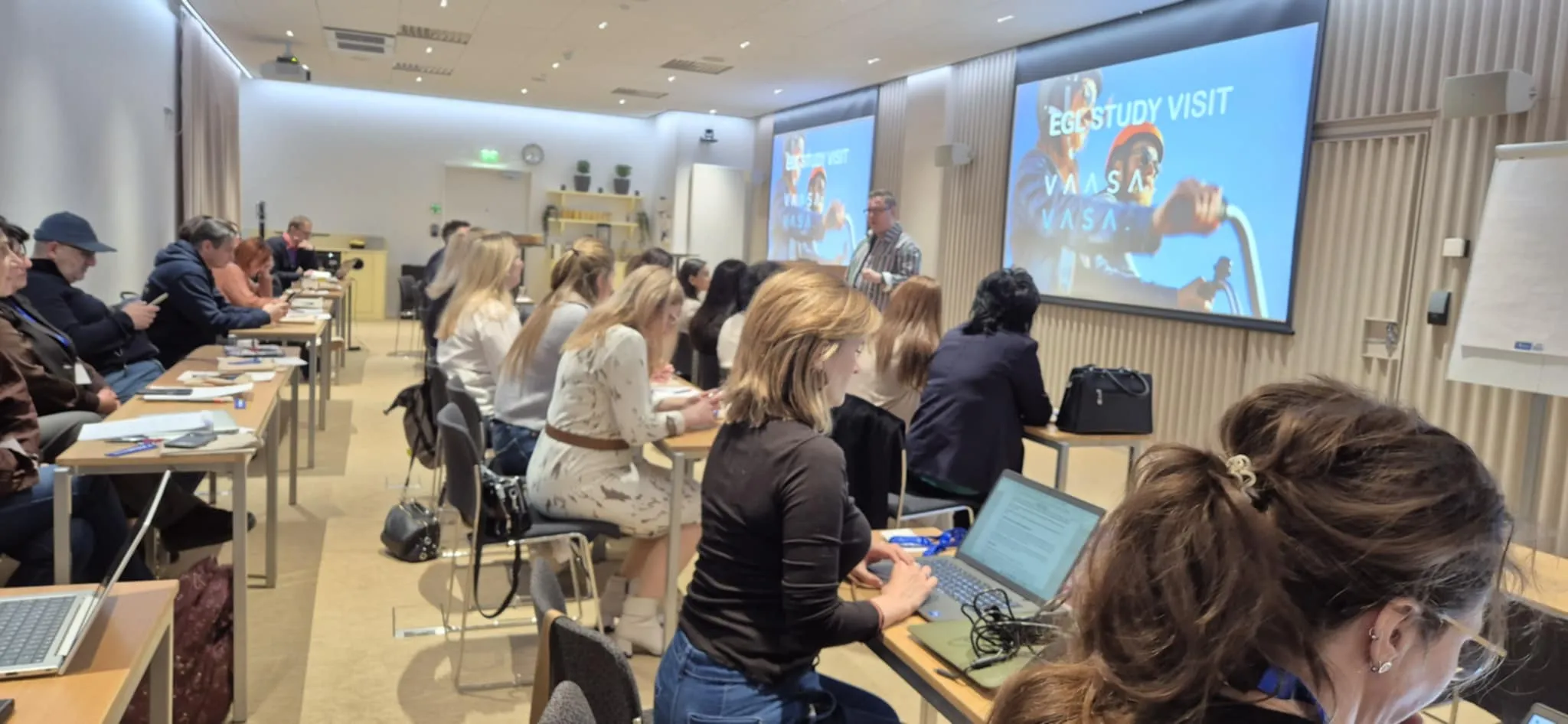 Sala konferencyjna wypełniona około dwudziestoma osobami siedzącymi przy długich stolikach. Uczestnicy korzystają z laptopów i notują. Mężczyzna stoi przy przodzie i prowadzi prezentację wyświetlaną na dwóch dużych ekranach z niebieskim tłem. Wnętrze ma jasne oświetlenie i drewniane panele na ścianie.