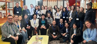 Około 28 osób stoi i siedzi wewnątrz w jasnym salonowym pomieszczeniu. Kilka osób siedzi na sofie i na podłodze, reszta stoi za nimi. W tle są drewniane regały z dekoracjami, jasne zasłony i czerwone panele; z przodu niski stół z wazonem.