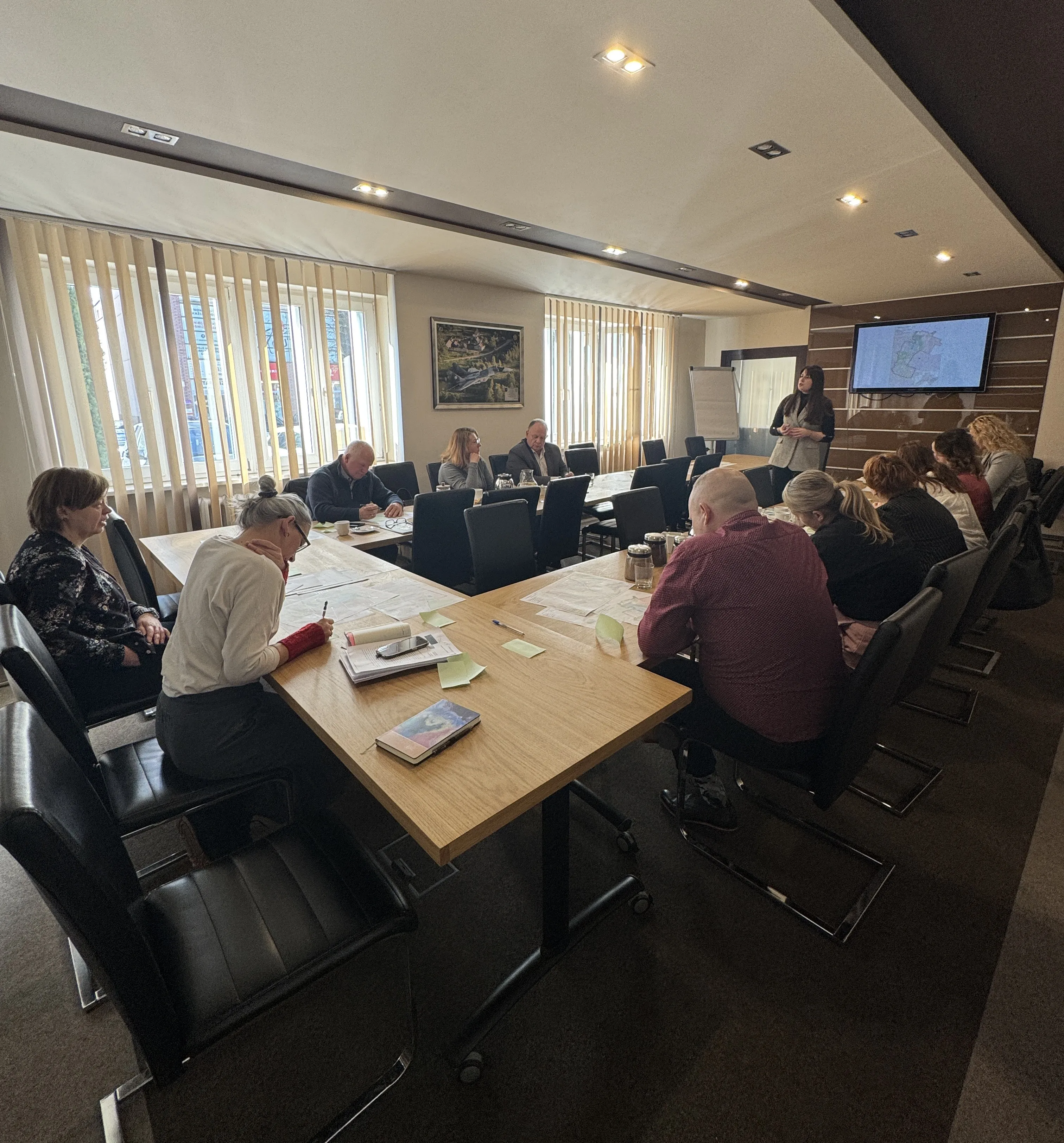 W sali konferencyjnej kilkanaście osób siedzi przy długim, drewnianym stole i przegląda dokumenty. Kobieta stoi przy froncie przy ekranie z mapą i prowadzi prezentację. Duże okna z pionowymi żaluzjami wpuszczają dzienne światło, na ścianie wisi obraz.