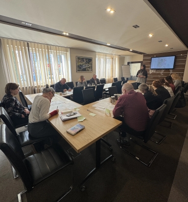 W sali konferencyjnej kilkanaście osób siedzi przy długim, drewnianym stole i przegląda dokumenty. Kobieta stoi przy froncie przy ekranie z mapą i prowadzi prezentację. Duże okna z pionowymi żaluzjami wpuszczają dzienne światło, na ścianie wisi obraz.