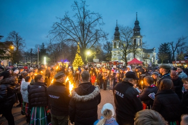 Grupa ludzi gromadzi się na placu wieczorem, trzymając płonące pochodnie. W tle widoczny kościół i oświetlona choinka. Niektórzy uczestnicy noszą tradycyjne stroje, dwie osoby mają ubrania z napisem "STRAŻ". Otoczenie wskazuje na zimową porę i świąteczną atmosferę.