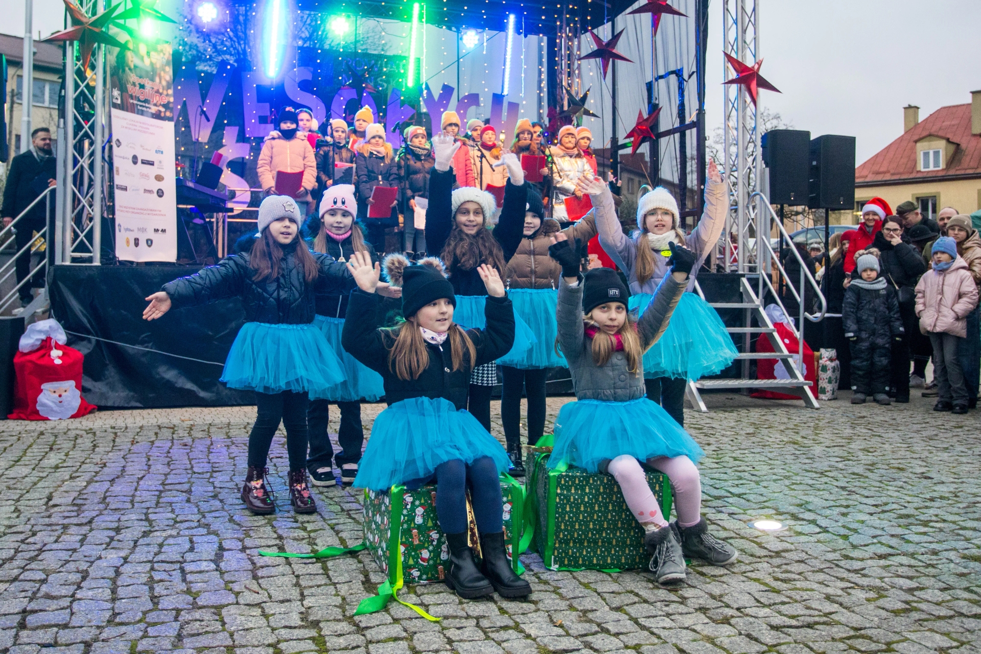 Grupa dzieci na zewnątrz podczas świątecznego występu, dziewczynki w niebieskich tiulowych spódniczkach, niektóre siedzą na dużych, ozdobnych prezentach, inne stoją z uniesionymi rękami, w tle scena z chórem dzieci i oświetleniem świątecznym, po prawej stronie widzowie ubrani na zimę.