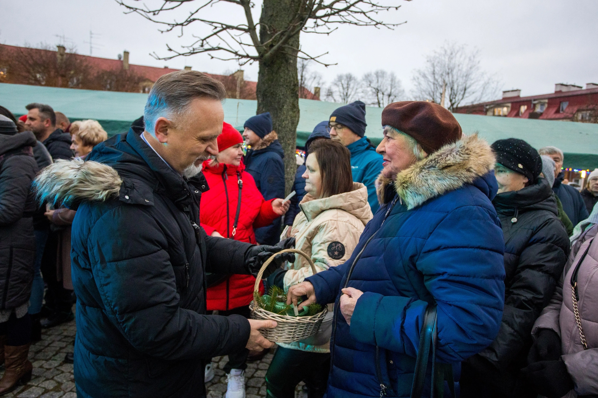 Grupa ludzi ubranych w zimowe ubrania stoi na zewnątrz przy drzewie, mężczyzna podaje kobiecie koszyk z gałązkami iglaków, w tle zielony namiot i domy.