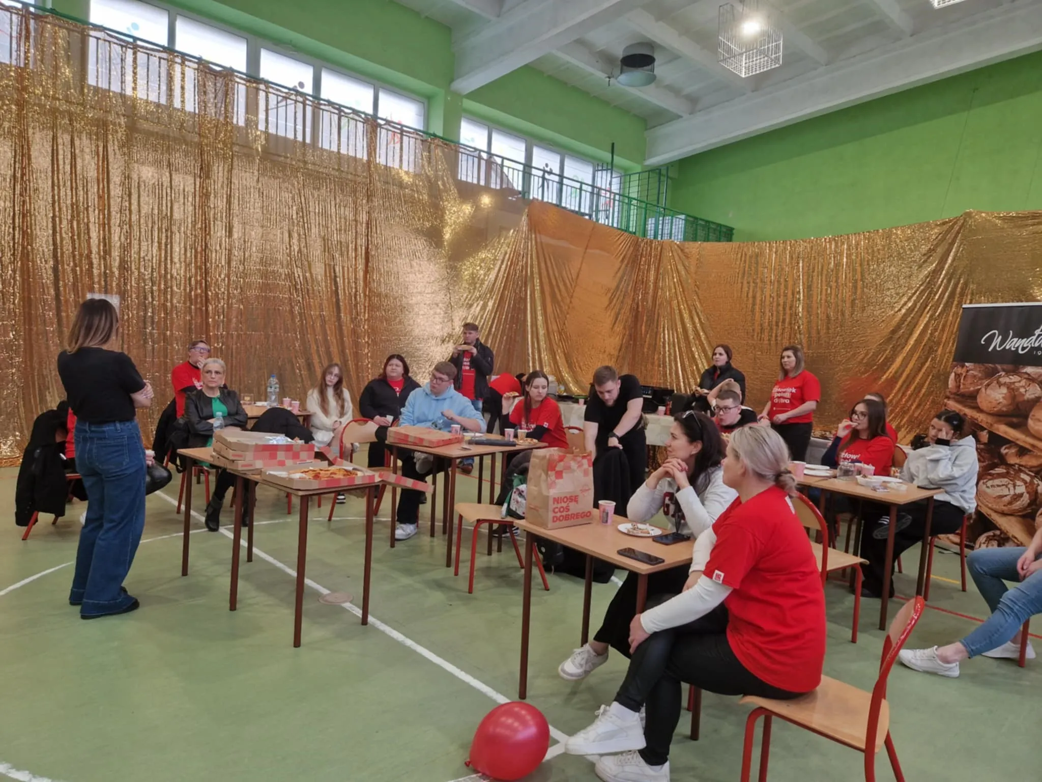 Grupa około piętnastu osób siedzi przy stolikach w sali gimnastycznej z zielonymi ścianami i dużymi oknami. Tło stanowi złota błyszcząca kotara. Na stolikach leżą pudła z pizzą i napoje. W większości osoby mają czerwone koszulki, jedna stoi zwrócona do grupy. Na podłodze leży czerwony balon.