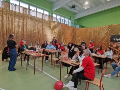 Grupa około piętnastu osób siedzi przy stolikach w sali gimnastycznej z zielonymi ścianami i dużymi oknami. Tło stanowi złota błyszcząca kotara. Na stolikach leżą pudła z pizzą i napoje. W większości osoby mają czerwone koszulki, jedna stoi zwrócona do grupy. Na podłodze leży czerwony balon.