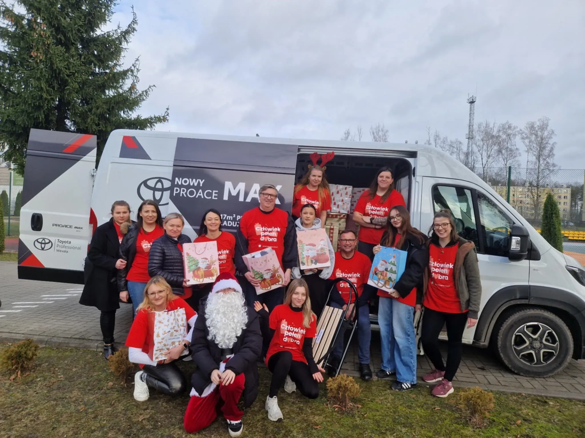 Grupa osób stoi i klęczy przed białym dostawczym samochodem na zewnątrz w ciągu dnia. Większość osób ma na sobie czerwone koszulki z białym napisem. Jedna osoba ubrana jest w strój Mikołaja z białą brodą. Kilka osób trzyma świąteczne torby na prezenty. W tle znajduje się szary niebo, drzewa i teren sportowy.