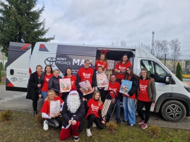 Grupa osób stoi i klęczy przed białym dostawczym samochodem na zewnątrz w ciągu dnia. Większość osób ma na sobie czerwone koszulki z białym napisem. Jedna osoba ubrana jest w strój Mikołaja z białą brodą. Kilka osób trzyma świąteczne torby na prezenty. W tle znajduje się szary niebo, drzewa i teren sportowy.