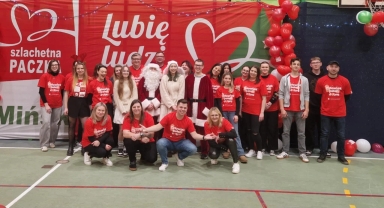 Grupa dwudziestu osób, z których większość ma czerwone koszulki z napisem, stoi i kuca na hali sportowej przed dużym czerwono-białym banerem z napisem „Szlachetna Paczka” i „Lubię ludzi”. W centrum dwie osoby przebrane za Świętego Mikołaja i kobietę w białym płaszczu oraz kilka osób w jasnych ubraniach. Po prawej stronie balony w kolorze czerwonym, zielonym i białym.
