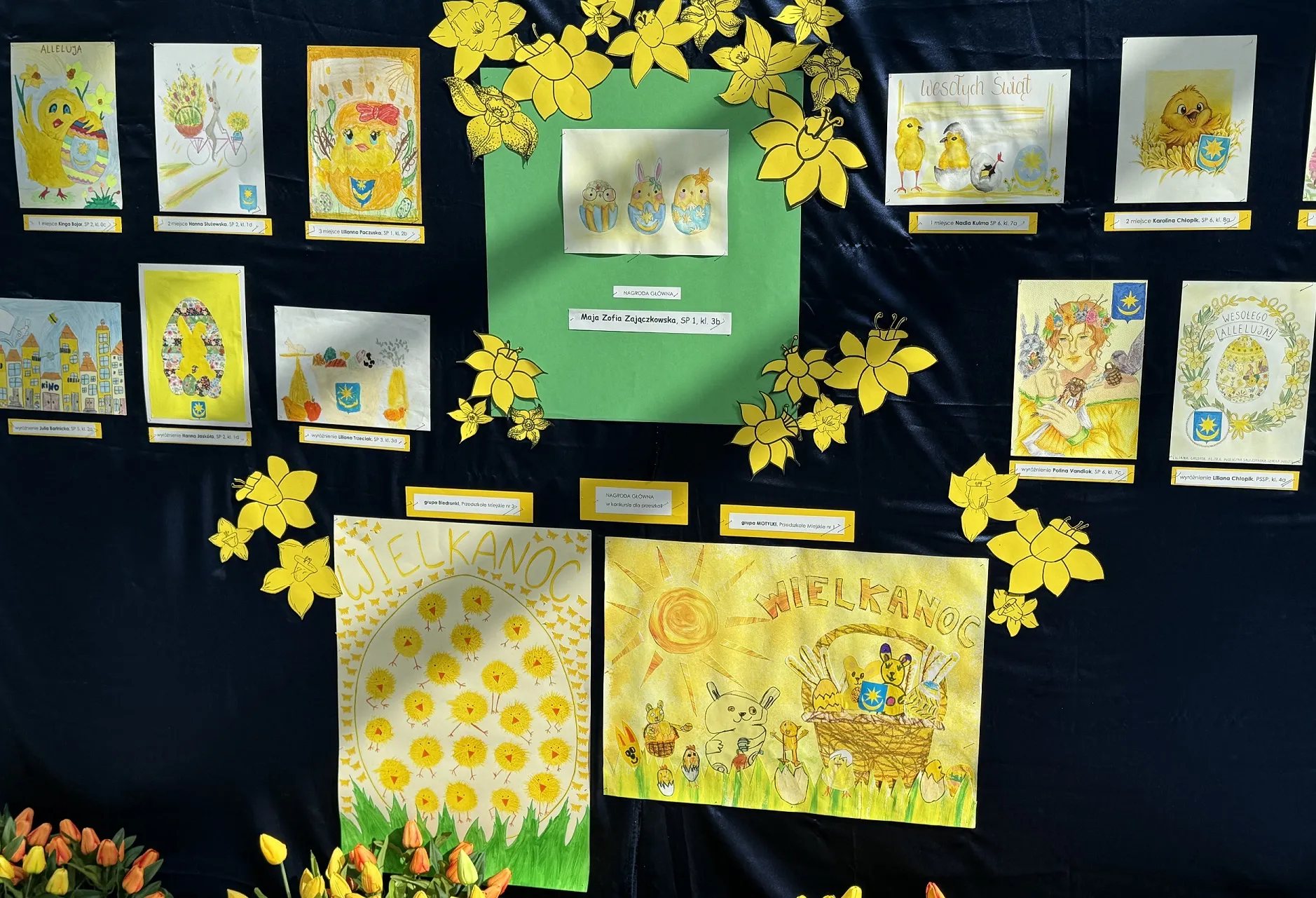 Tablica wystawowa z kilkunastoma dziecięcymi rysunkami wielkanocnymi na ciemnym tle. Na środku zielona ramka z rysunkiem trzech kurcząt, wokół papierowe żonkile, u dołu dwa większe plakaty z motywami jajek, koszyków i kurcząt oraz bukiety tulipanów.