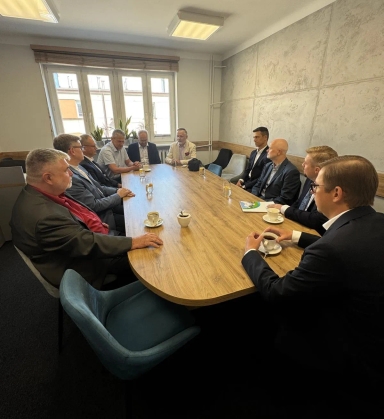 Dziesięciu mężczyzn w garniturach siedzi przy dużym drewnianym stole w sali konferencyjnej, rozmawiając i pijąc kawę.