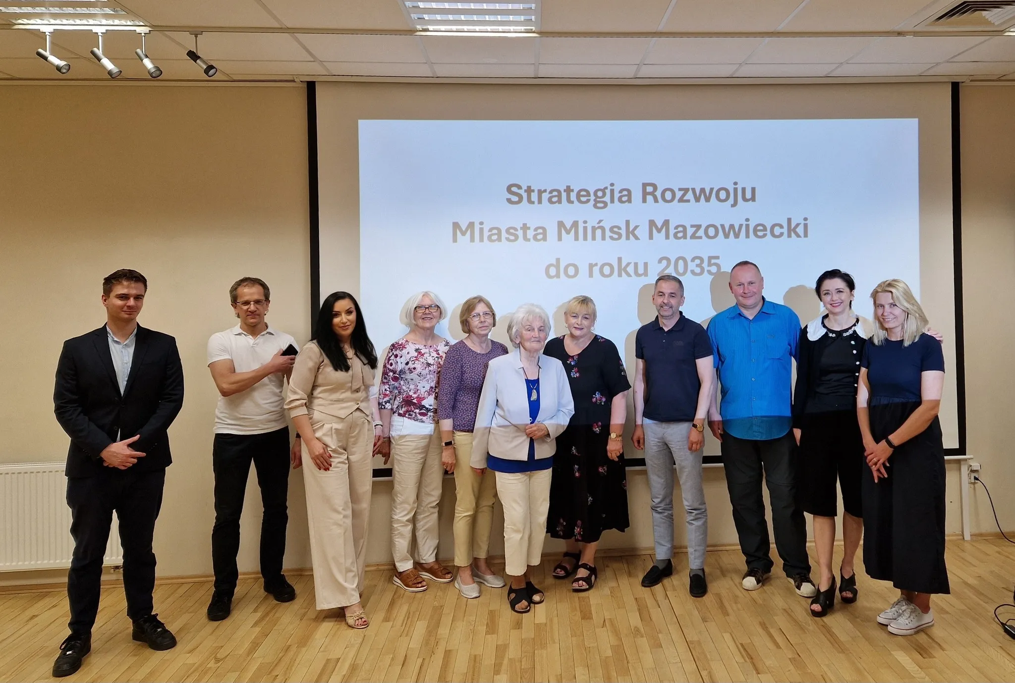 Na zdjęciu stoi dziesięć osób w różnych strojach w sali z jasnym drewnianym podłogą, z prezentacją na ścianie w tle, dotyczącą strategii rozwoju Miasta Mińsk Mazowiecki do roku 2035.
