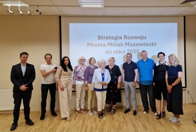 Na zdjęciu stoi dziesięć osób w różnych strojach w sali z jasnym drewnianym podłogą, z prezentacją na ścianie w tle, dotyczącą strategii rozwoju Miasta Mińsk Mazowiecki do roku 2035.