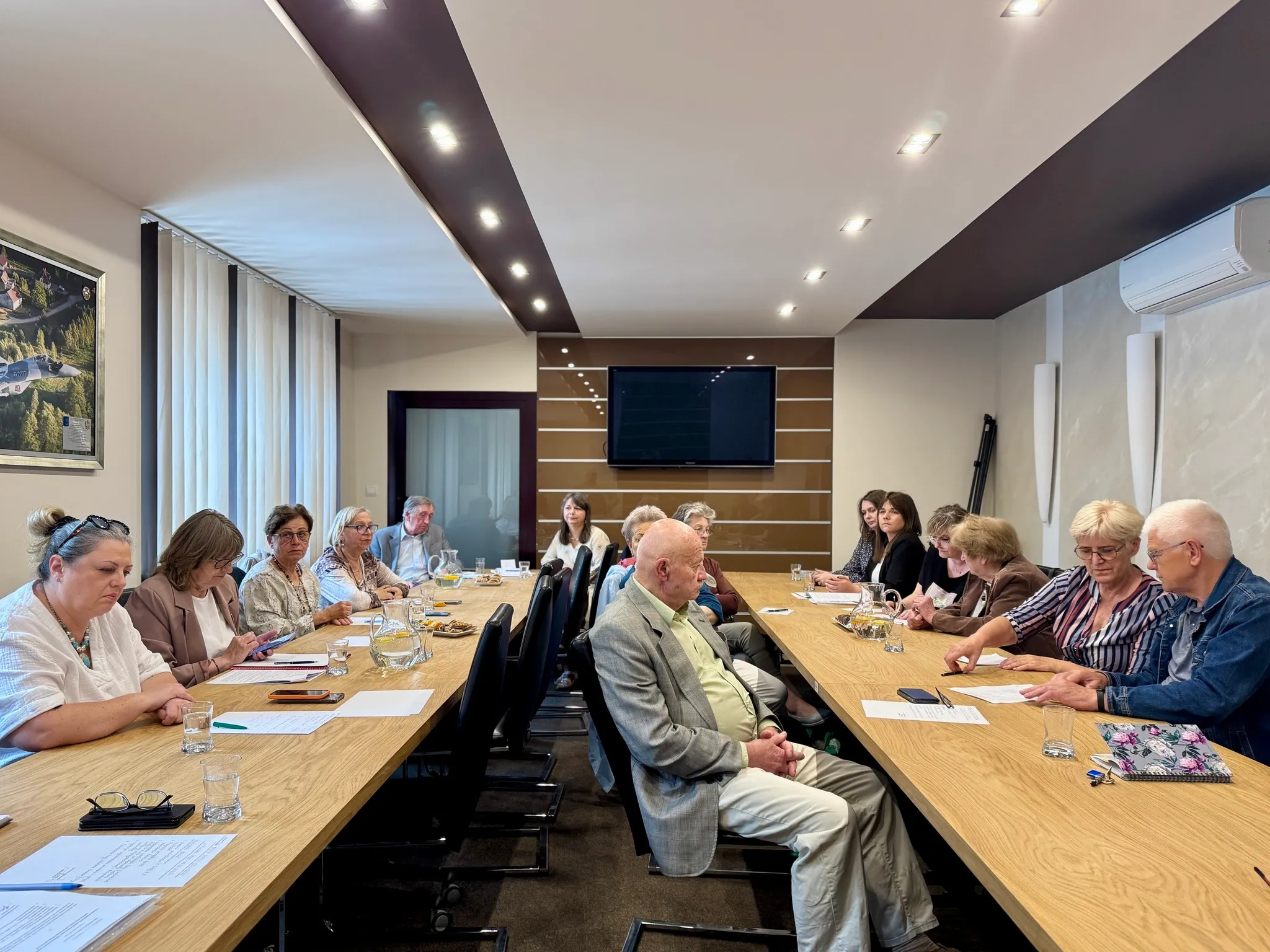 W sali konferencyjnej przy długim stole siedzi grupa dziesięciu osób, rozmawiają o dokumentach i używają telefonów, w tle widoczne okno z zasłonami oraz ekran.