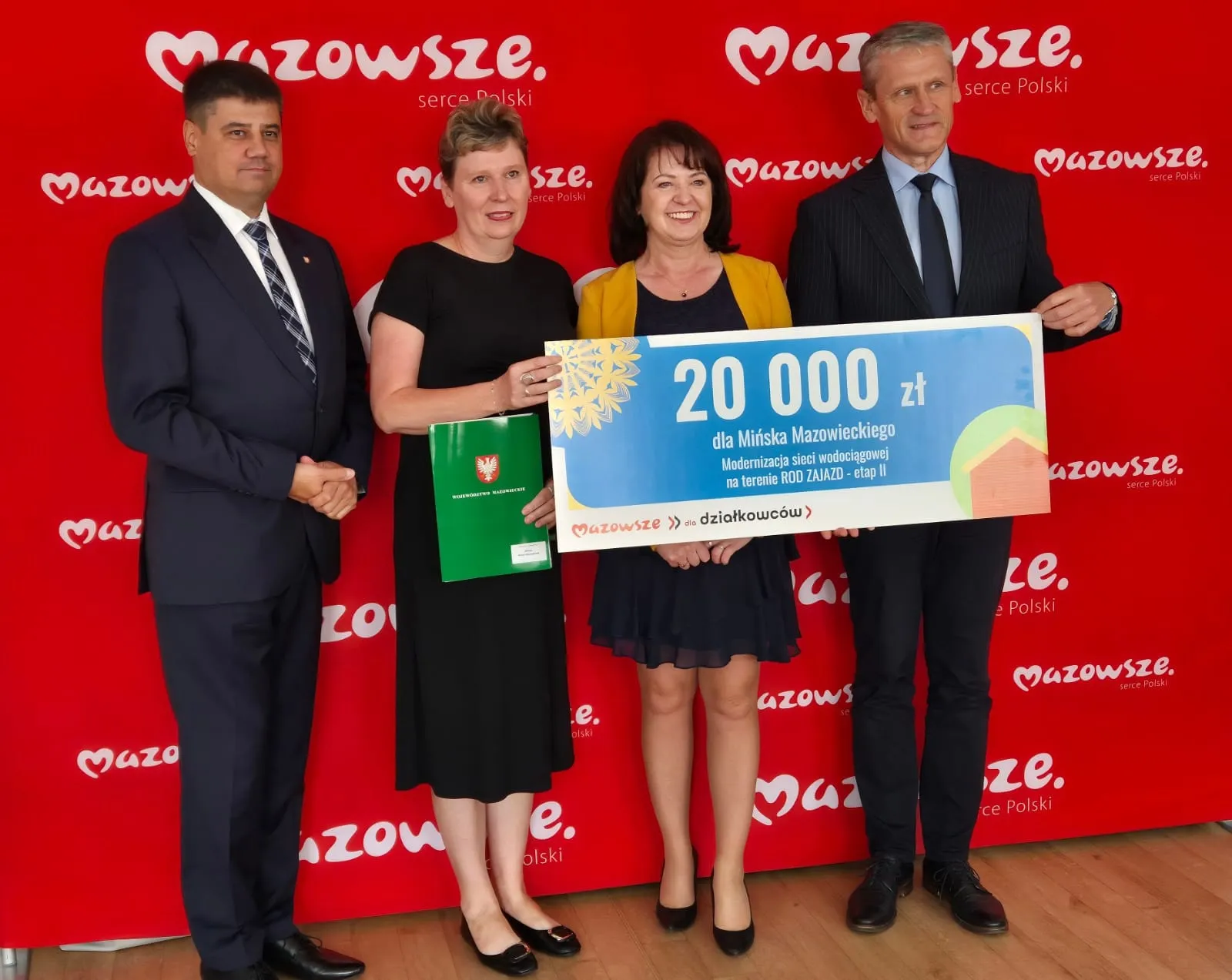 Cztery osoby stoją razem na tle czerwonego baneru z napisem "Mazowsze, serce Polski", trzymając duży czek na kwotę 20 000 zł oraz dokumenty związane z projektem modernizacji sieci wodociągowej.