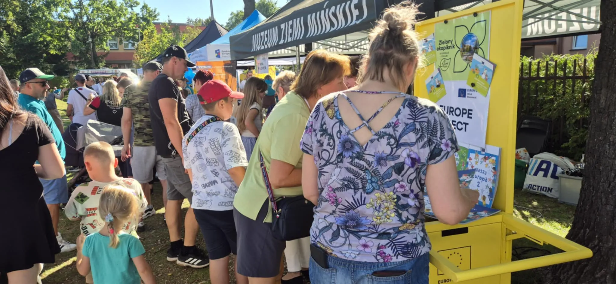 Grupa ludzi, w tym dzieci i dorośli, stoi przy żółtym stoisku na zewnątrz, przy słonecznej pogodzie, w parku z drzewami i namiotami wystawowymi z napisem „Muzeum Ziemi Mińskiej”.