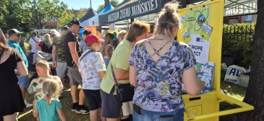 Grupa ludzi, w tym dzieci i dorośli, stoi przy żółtym stoisku na zewnątrz, przy słonecznej pogodzie, w parku z drzewami i namiotami wystawowymi z napisem „Muzeum Ziemi Mińskiej”.