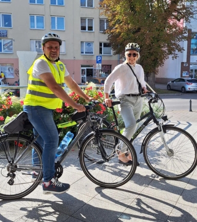 Dwóch dorosłych na rowerach stoi na chodniku przy nasadzeniu kwiatów w miejskiej okolicy w ciągu dnia. Mężczyzna po lewej ma na sobie kask i żółtą odblaskową kamizelkę, kobieta po prawej nosi kask, białą bluzkę i jasne spodnie. W tle budynki mieszkalne, samochody i drzewa.