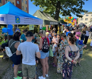 Grupa dorosłych i dzieci stoi przed namiotami z napisami podczas plenerowego wydarzenia w dzień.