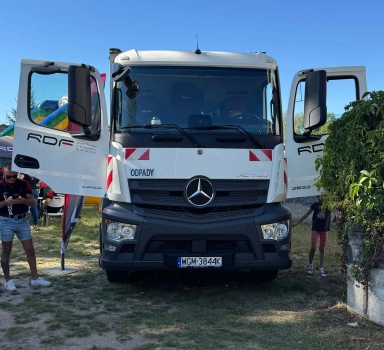 Ciężarówka Mercedes z otwartymi drzwiami stoi na trawie w dziennym świetle, obok po lewej stoi mężczyzna, a po prawej dziecko przy krzewie.