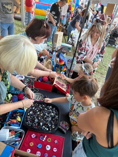Dziewczynka i chłopiec stoją przy stoisku z guzikami i materiałami do rękodzieła, a trzy dorosłe kobiety im pomagają podczas imprezy plenerowej w ciągu dnia.