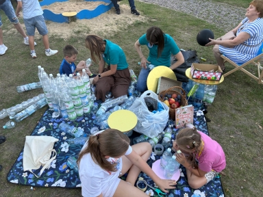 Cztery osoby, w tym dwoje dzieci i dwie kobiety siedzą na kocu na trawie na zewnątrz, otoczone wieloma plastikowymi butelkami i owocami, obok kobieta siedzi na składanym krześle trzymając czarną piłkę.